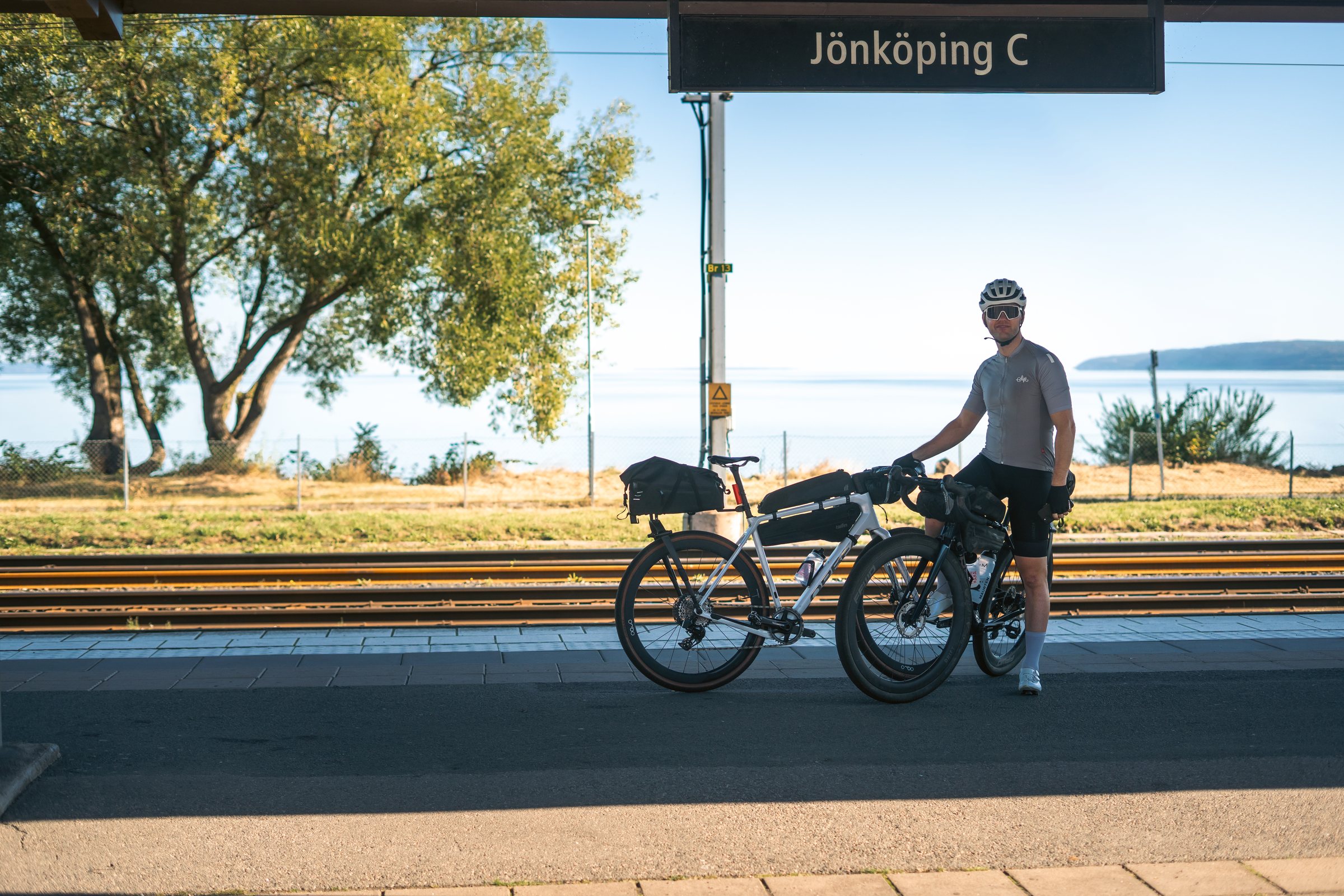 Cyclist with two loaded gravel bikes under the Jönköping C sign, Lake Vättern in the background