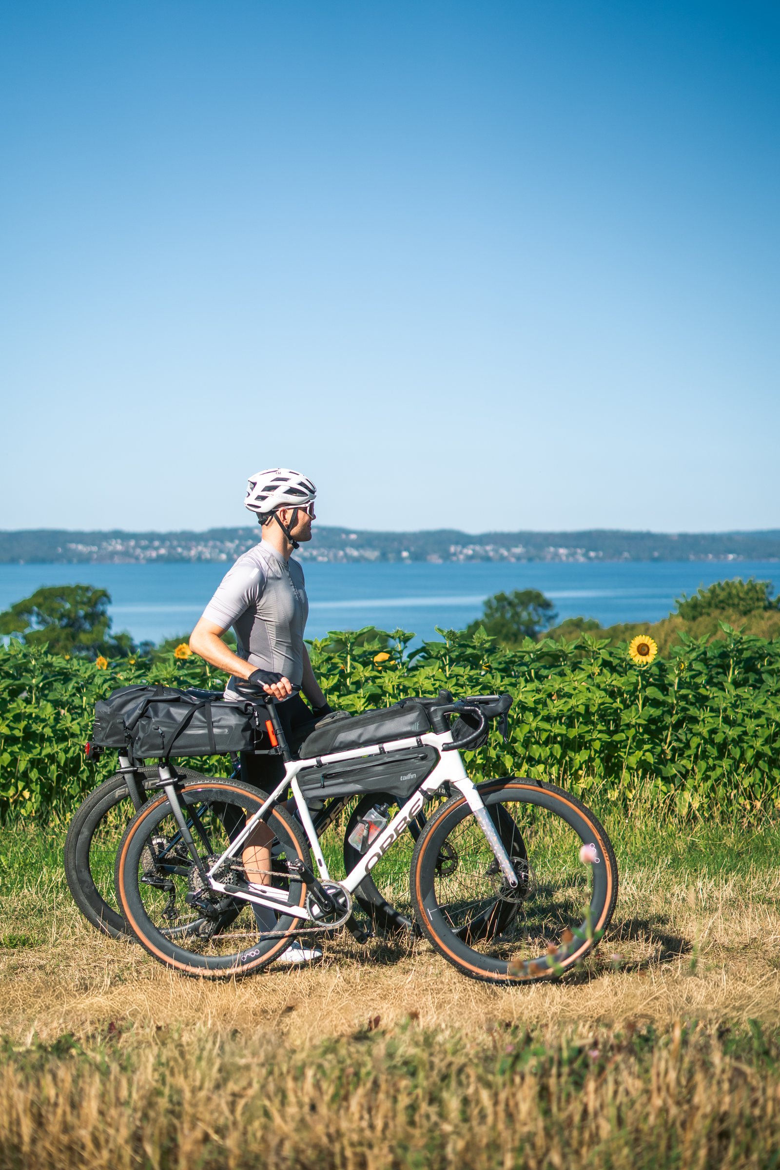 Cyclist with a loaded gravel bike at the edge of a sunflower field, Lake Vättern behind