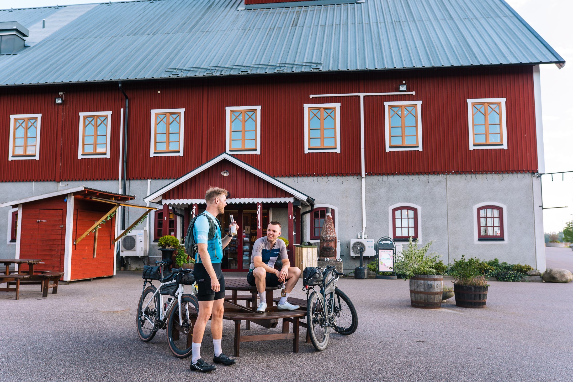 Two cyclists at a picnic table outside the red Brunneby Musteri building
