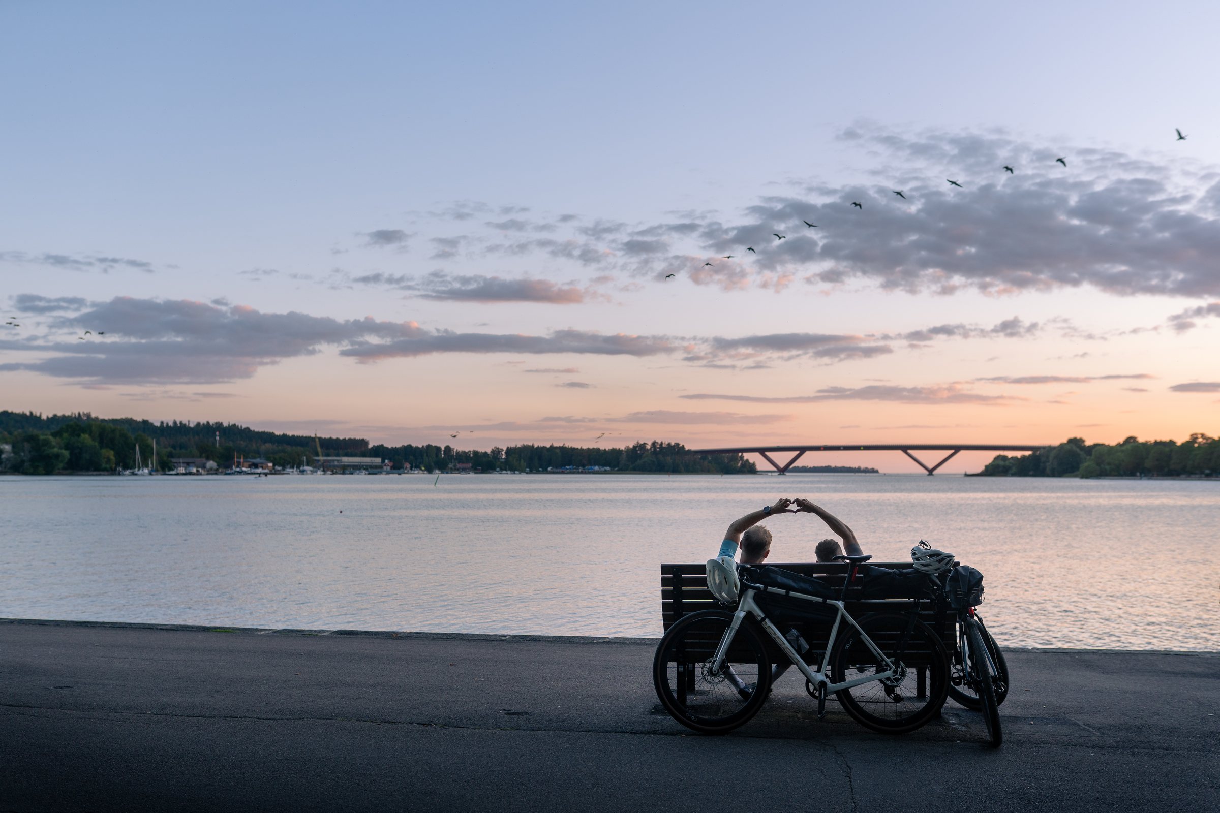 Two cyclists on a bench by Lake Vättern in Motala at sunset, bikes leaning beside them